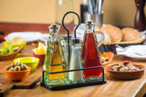 A TableCraft Marbella oil & vinegar set sits in a metal caddy on a wooden table, surrounded by bowls of olives, lemon wedges, salad, bread, and peppers—evoking a Mediterranean meal setting with cutlery in the background.