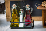 Three glass bottles in a black TableCraft Marbella holder: one with yellow olive oil, one with dark red vinegar, and a silver salt shaker in the center—this elegant oil & vinegar set rests on a white tablecloth against a wooden background.