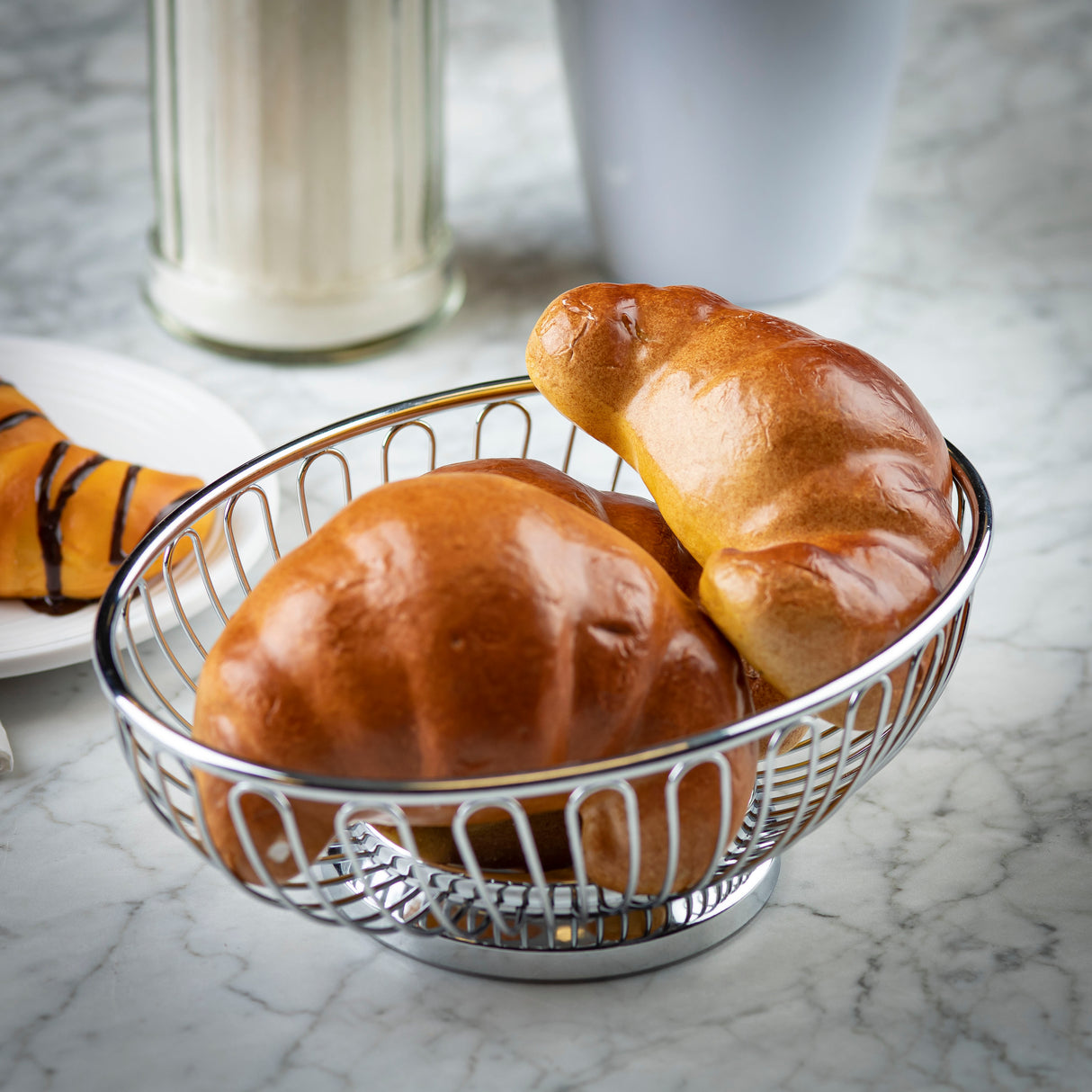 A stainless steel basket holds two shiny, golden-brown croissants on a marble surface. In the background, there’s a chocolate-drizzled pastry, a glass sugar dispenser, and a white cup, all slightly out of focus.