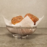 A stainless steel basket lined with white parchment paper holds two everything bagels topped with assorted seeds, sitting on a gray stone countertop against a plain gray wall.