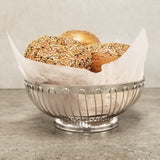 A stainless steel Regent Basket by TableCraft Products, lined with parchment paper, holds three round, golden-brown bread rolls topped with mixed seeds on a smooth gray surface against a plain light gray background.