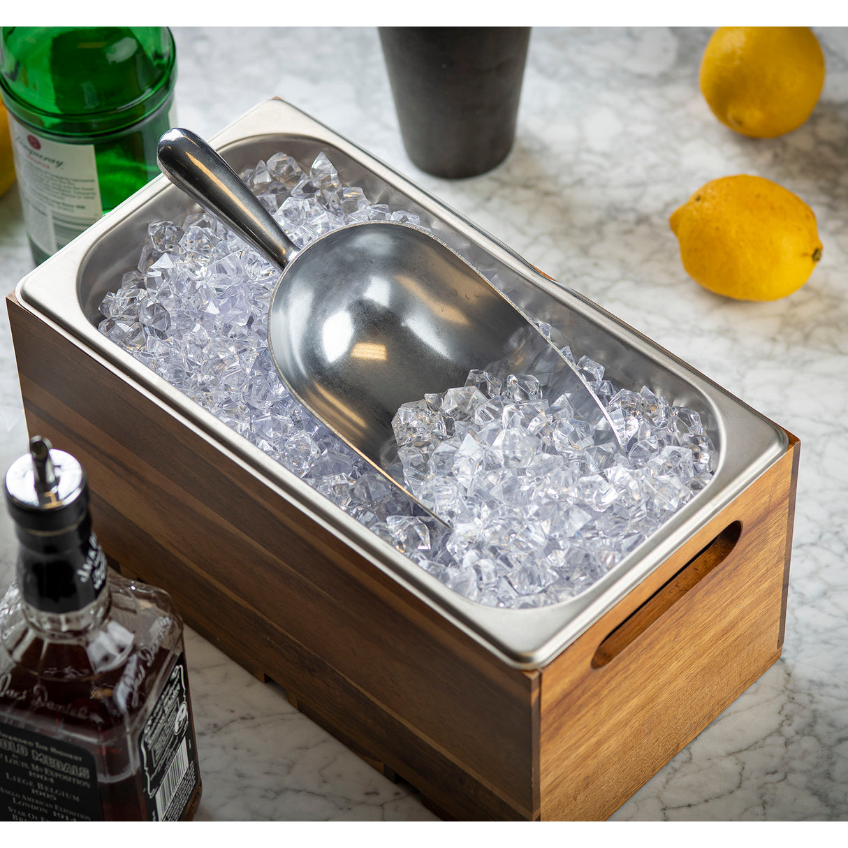 An aluminum ice scoop rests on clear ice cubes in a rectangular 24 oz. stainless steel tray with a wooden outer frame, placed on a marble countertop. Bottles and whole lemons are visible in the background.