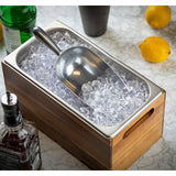 An aluminum ice scoop rests on clear ice cubes in a rectangular 24 oz. stainless steel tray with a wooden outer frame, placed on a marble countertop. Bottles and whole lemons are visible in the background.