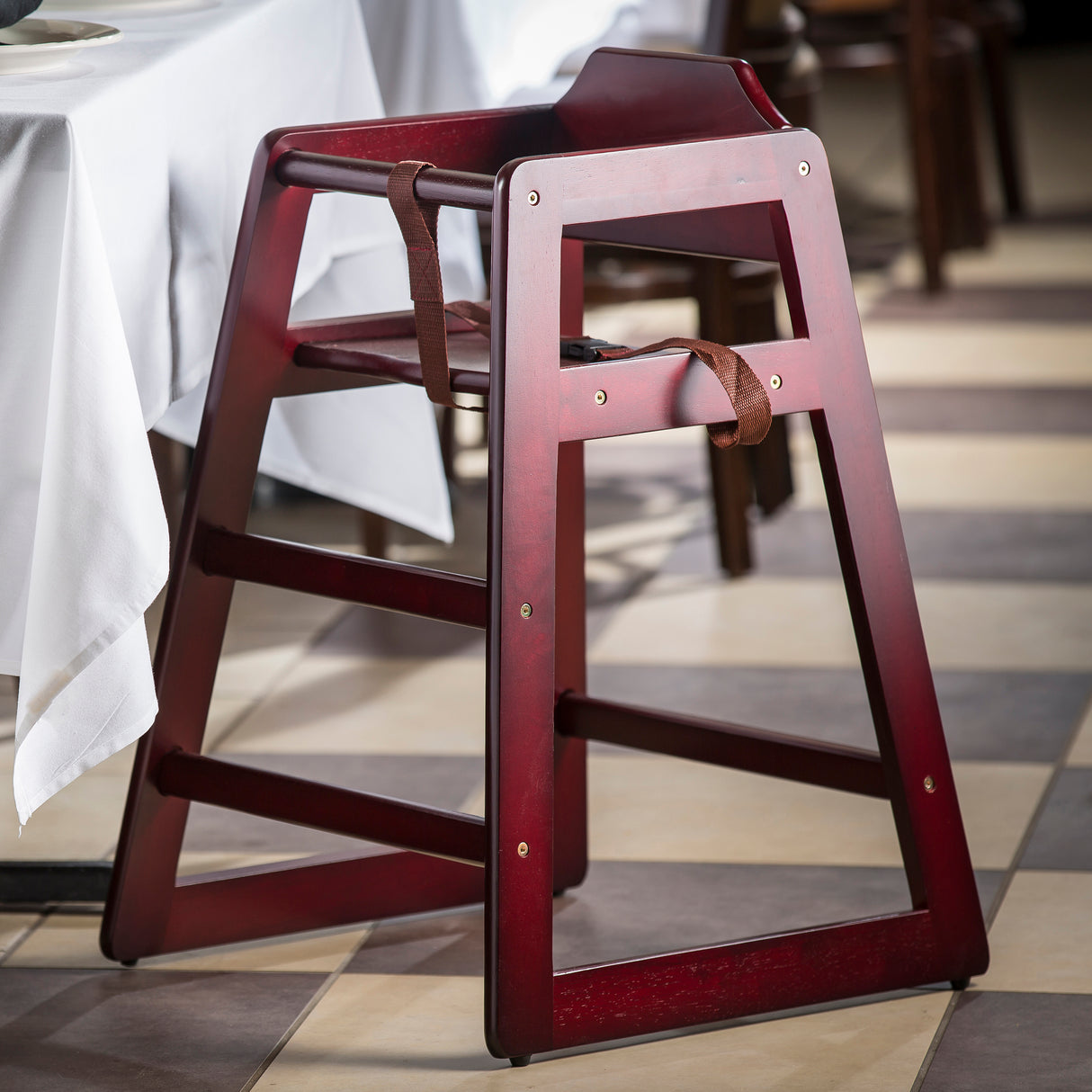 A stackable wooden restaurant high chair with a dark red finish and a brown safety strap sits next to a table with a white tablecloth. The empty rubberwood chair rests on a checkered tile floor in beige and brown tones.