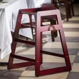 A stackable wooden restaurant high chair with a dark red finish and a brown safety strap sits next to a table with a white tablecloth. The empty rubberwood chair rests on a checkered tile floor in beige and brown tones.
