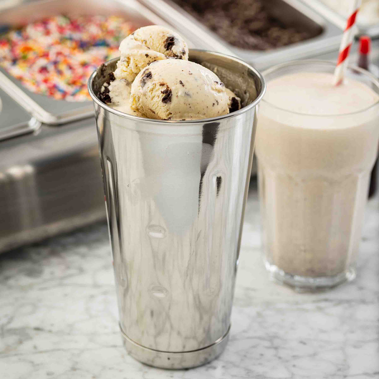 A 30 oz stainless steel malt cup filled with scoops of cookie dough ice cream sits on a marble counter. Behind it is a glass with a creamy milkshake and red-striped straw, surrounded by sprinkles and toppings in metal trays.