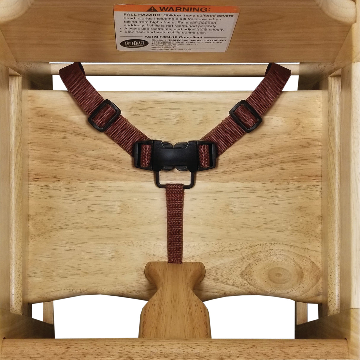 A top-down view of a stackable wooden high chair with a dark red safety harness and plastic buckle. A wooden crotch post separates the leg openings. Features a white warning label and natural wood finish; ASTM F404-18 compliant.