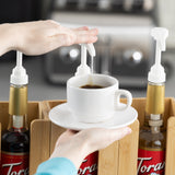 A person presses a 1/4 oz syrup pump on a syrup bottle, dispensing syrup into a white cup of coffee on a saucer. Two similar dishwasher safe bottles sit in a wooden organizer, with a blurred kitchen appliance in the background.
