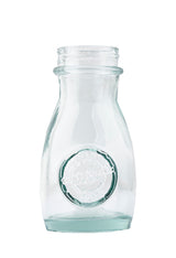 A clear, empty, vintage TableCraft 6618 glass milk bottle with a round embossed logo on the front. The bottle has a short neck, flared rim, and slightly curved sides. The plain white background emphasizes its transparent and clean appearance.