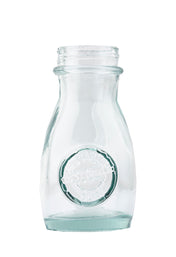 A clear, empty, vintage TableCraft 6618 glass milk bottle with a round embossed logo on the front. The bottle has a short neck, flared rim, and slightly curved sides. The plain white background emphasizes its transparent and clean appearance.