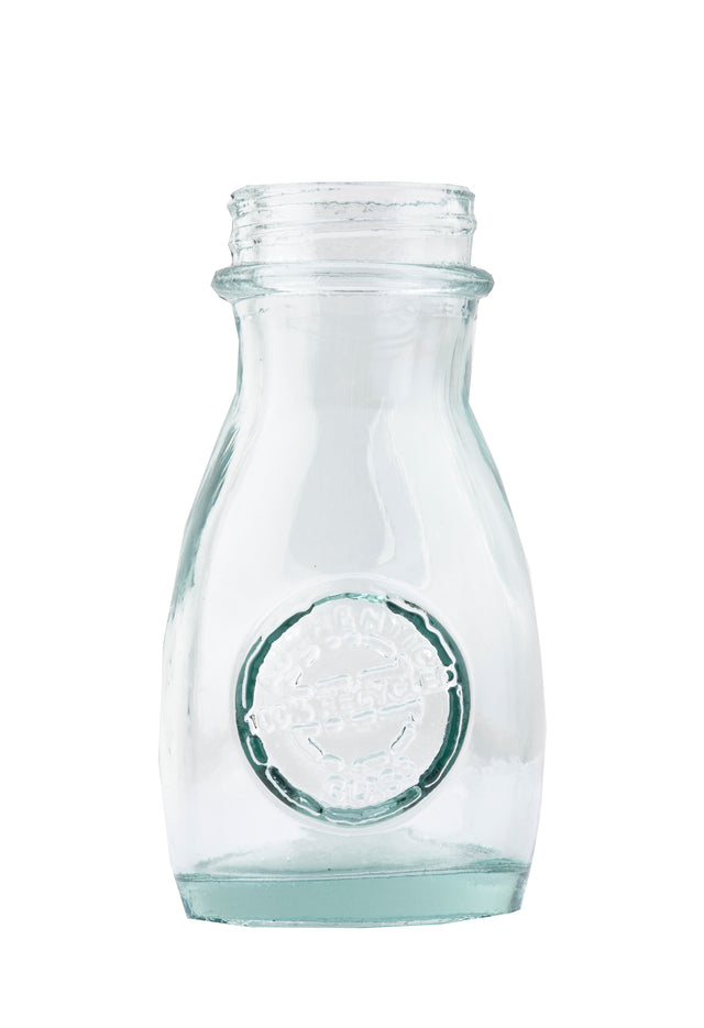A clear, empty, vintage TableCraft 6618 glass milk bottle with a round embossed logo on the front. The bottle has a short neck, flared rim, and slightly curved sides. The plain white background emphasizes its transparent and clean appearance.