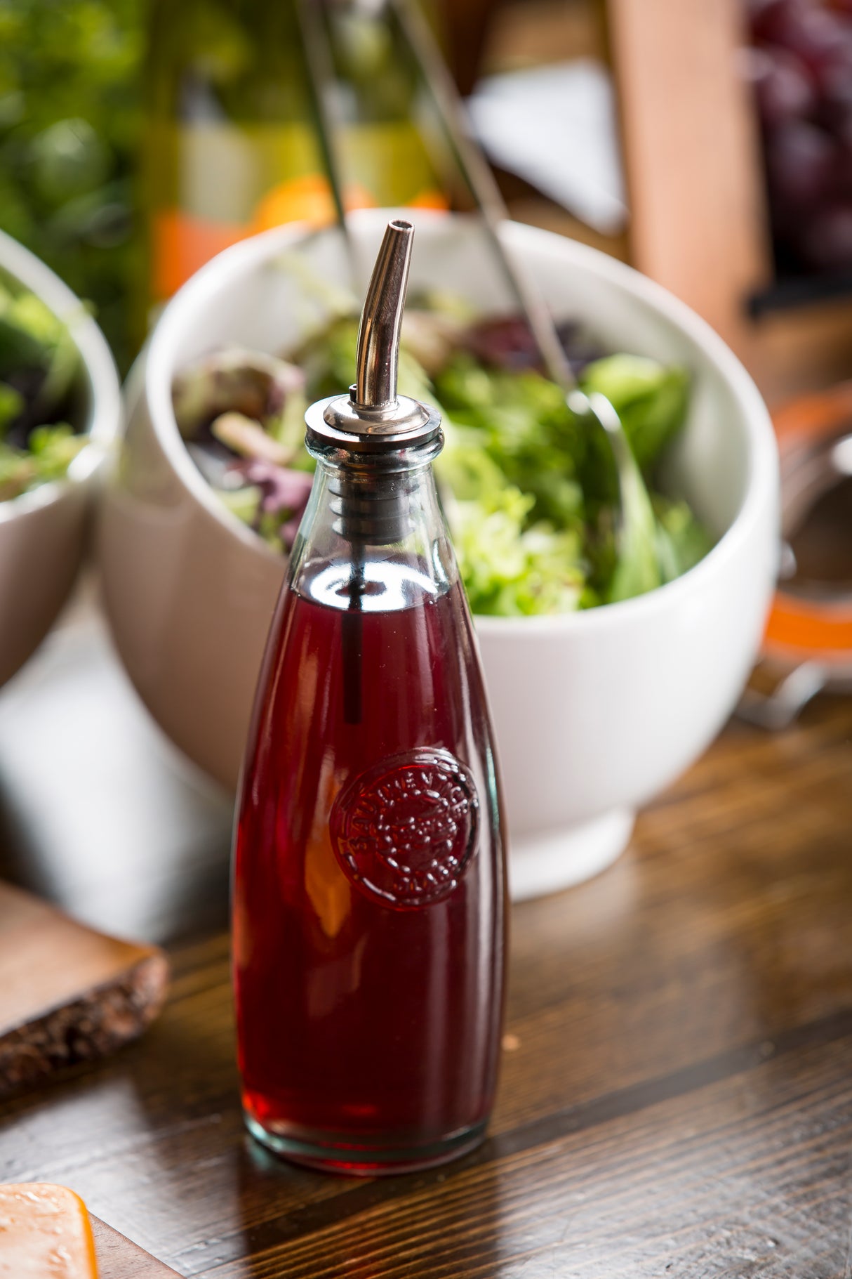 A recycled glass bottle filled with red vinaigrette sits on a wooden table in front of a salad bowl. Featuring a stainless steel top, this Oil & Vinegar Dispenser stands amid additional bowls and blurred food items in the background.