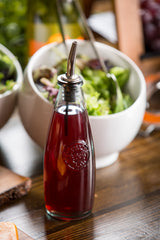 A recycled glass bottle filled with red vinaigrette sits on a wooden table in front of a salad bowl. Featuring a stainless steel top, this Oil & Vinegar Dispenser stands amid additional bowls and blurred food items in the background.