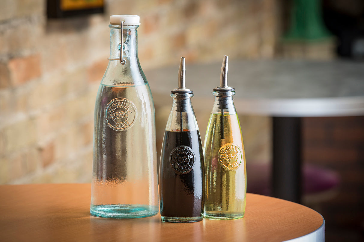 Three recycled glass bottles sit on a wooden table: a tall water bottle with a clasp lid, and two Oil & Vinegar Dispensers with stainless steel tops—one filled with dark balsamic vinegar, the other yellow olive oil. A blurred brick wall is in the background.