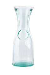 A tall, clear authentic carafe with a narrow neck and wide mouth. Crafted from recycled glass with a subtle green tint, it holds 28 oz. The slightly thicker bottom and embossed circular seal add elegance against a plain white background.