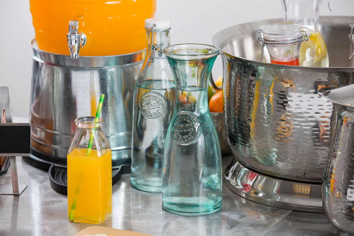 An authentic carafe made from recycled glass holds orange juice with a green straw, sitting on a metal counter beside two clear 28 oz bottles, a beverage dispenser with orange drink, and an ice bucket filled with bottled fruit drinks.