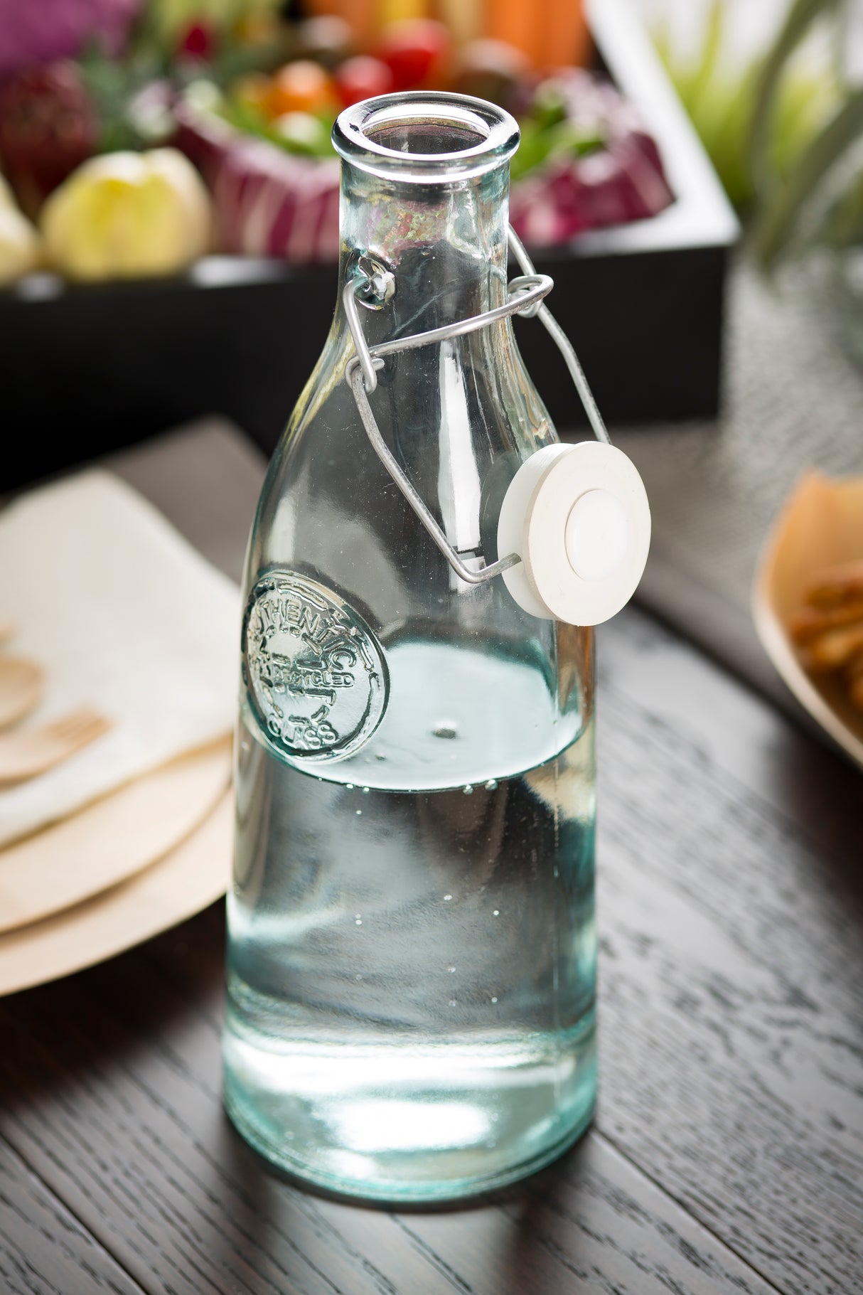 A clear 33 oz recycled glass bottle with a ceramic swing top is filled with water and placed on a dark wooden table. Blurred food items and colorful vegetables in the background create a fresh, inviting atmosphere.