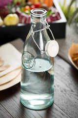 A clear 33 oz recycled glass bottle with a ceramic swing top is filled with water and placed on a dark wooden table. Blurred food items and colorful vegetables in the background create a fresh, inviting atmosphere.