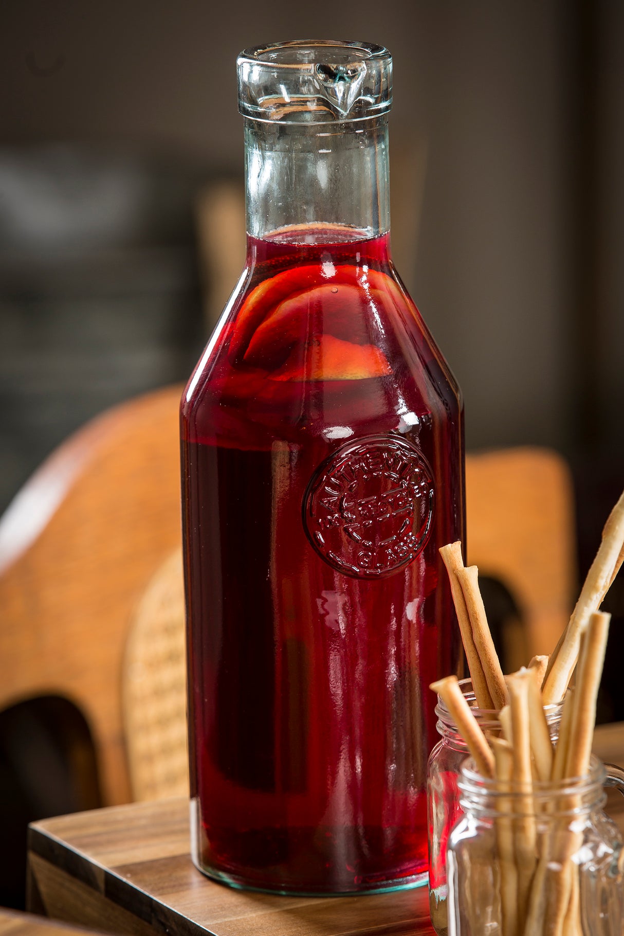 A clear apothecary bottle with a cork stopper, filled with deep red liquid and visible fruit slices, likely sangria, sits on a wooden surface. Next to it, a jar holds several thin breadsticks. The softly blurred background shows wooden chairs.