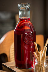 A clear apothecary bottle with a cork stopper, filled with deep red liquid and visible fruit slices, likely sangria, sits on a wooden surface. Next to it, a jar holds several thin breadsticks. The softly blurred background shows wooden chairs.