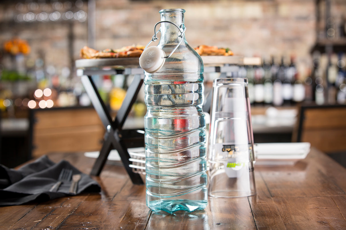 A clear, ribbed recycled glass water bottle with a latch top sits on a wooden table. Two clear cups are stacked upside-down beside it. In the blurred background, shelves of wine bottles create a cozy restaurant scene.