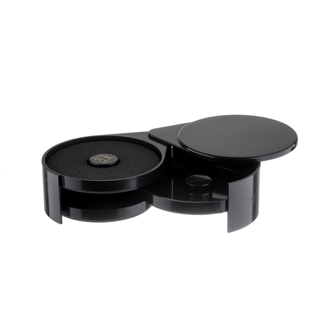 A glossy black, circular container with two sections; one features a swing-out tray revealing a foam pad and a coin cell battery, while the other stays closed with a flat lid. The sleek, modern design boasts smooth, shiny surfaces.