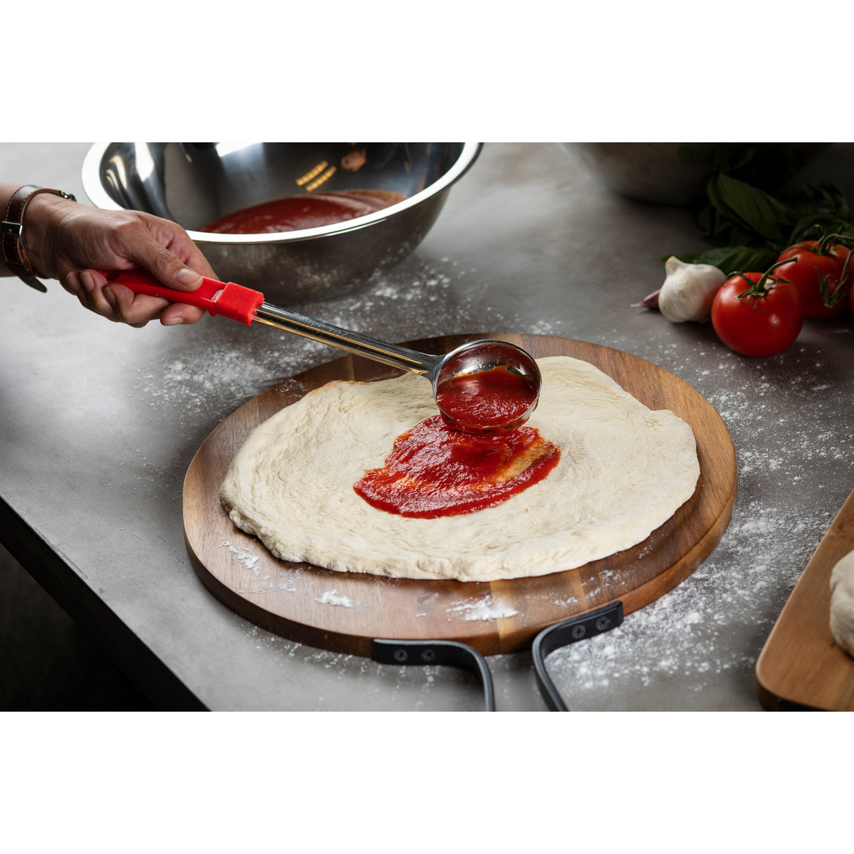 A hand uses a spoonout with a red handle to spread tomato sauce on raw pizza dough atop a wooden pizza peel. Nearby are fresh tomatoes, garlic, basil, a stainless steel mixing bowl, and scattered flour on a gray countertop.