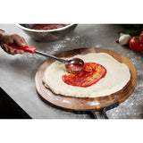 A hand uses a stainless steel ladle with a red handle to spread tomato sauce onto round pizza dough on a wooden board. Flour is scattered on the gray countertop, and in the background are fresh tomatoes, garlic, basil, and a metal bowl of sauce.