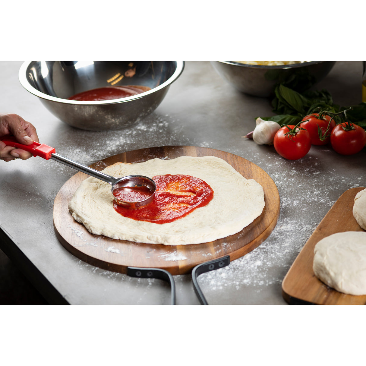 A hand spreads tomato sauce on raw pizza dough with a stainless steel ladle. The dough sits on a wooden pizza peel dusted with flour. Nearby are metal bowls, fresh tomatoes, basil, garlic, and extra dough balls on a floured countertop.