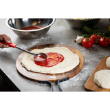 A hand spreads tomato sauce on raw pizza dough with a stainless steel ladle. The dough sits on a wooden pizza peel dusted with flour. Nearby are metal bowls, fresh tomatoes, basil, garlic, and extra dough balls on a floured countertop.