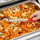 A hand with a spoonout featuring a red handle is scooping mixed vegetables—carrots, corn, peas, green beans, and diced potatoes—from a stainless steel buffet tray. The vibrant vegetables are cut into small cubes.