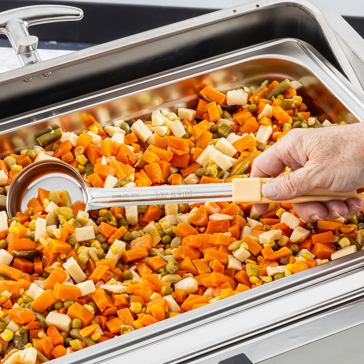 A hand holding a Spoonout stainless steel serving ladle hovers over a tray filled with freshly cooked, cubed carrots, parsnips, and green beans. The evenly cut vegetables look vibrant and ready to be enjoyed. The ladle is dishwasher safe.