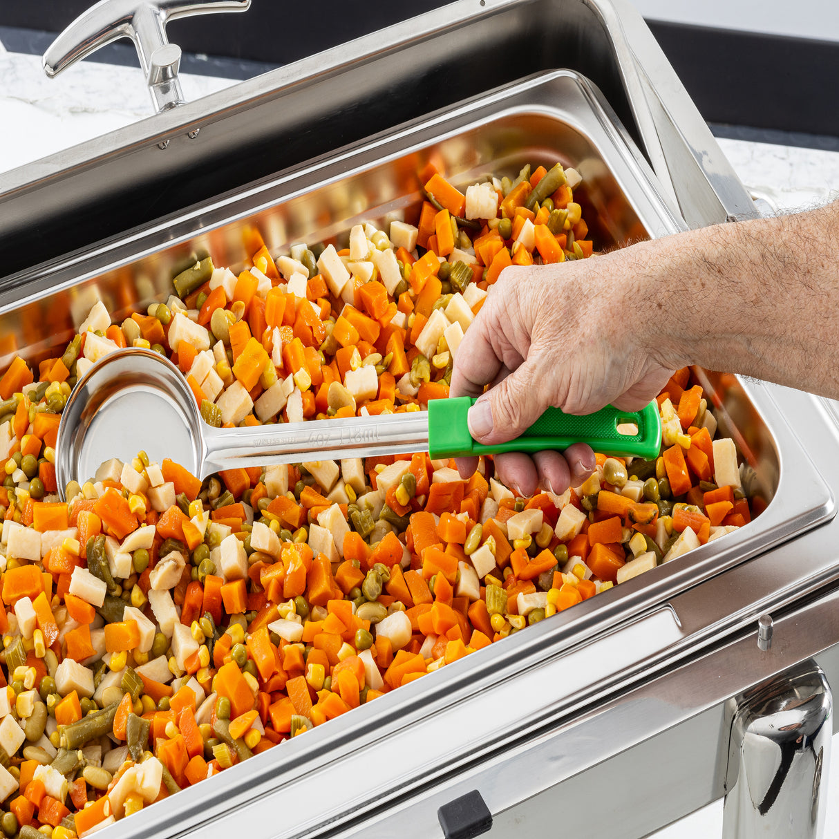 A hand uses a large Spoonout stainless steel ladle with a green handle to serve a colorful mix of vegetables from a tray, likely part of a buffet or cafeteria setup. The ladle is durable and dishwasher safe for easy cleaning.