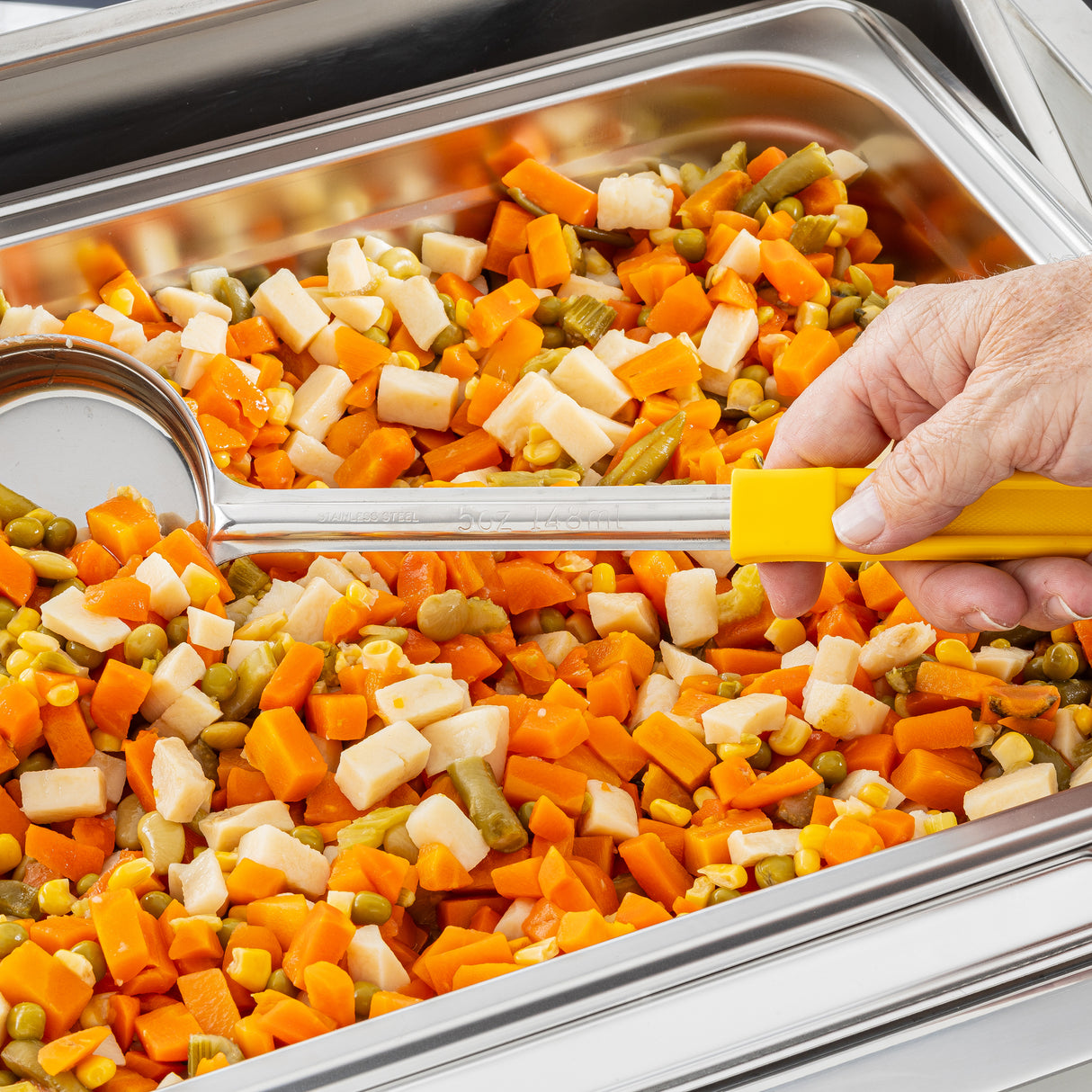 A hand holding a yellow-handled Spoonout serving spoon scoops mixed diced vegetables—carrots, green beans, peas, corn, and potatoes—from a stainless steel food tray. The colorful vegetables are evenly mixed and freshly cooked.