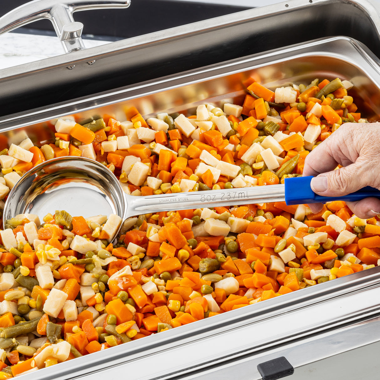A hand holds a large Spoonout silver serving spoon with a blue handle, scooping a colorful mix of diced carrots, potatoes, green beans, and peas from a stainless steel buffet tray. The dishwasher safe tray is filled to the brim with cooked vegetables.