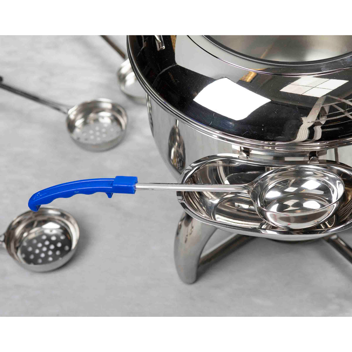 A shiny stainless steel fondue pot with a reflective surface sits on a stand. Several dishwasher safe, slotted and solid fondue forks with blue handles are nearby, one resting on a side tray attached to the pot. The background is light gray.