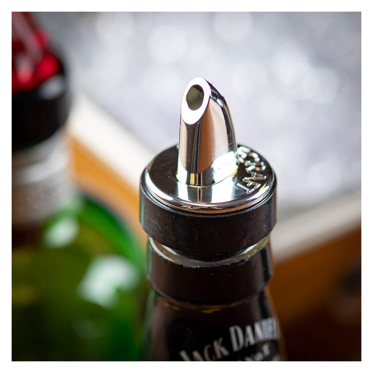 Close-up of a chrome plated Free Flow Pourer on a Jack Daniel’s bottle. The shiny, BPA free spout is centered in the frame, with softly blurred bottles and a bright, bokeh background adding depth.