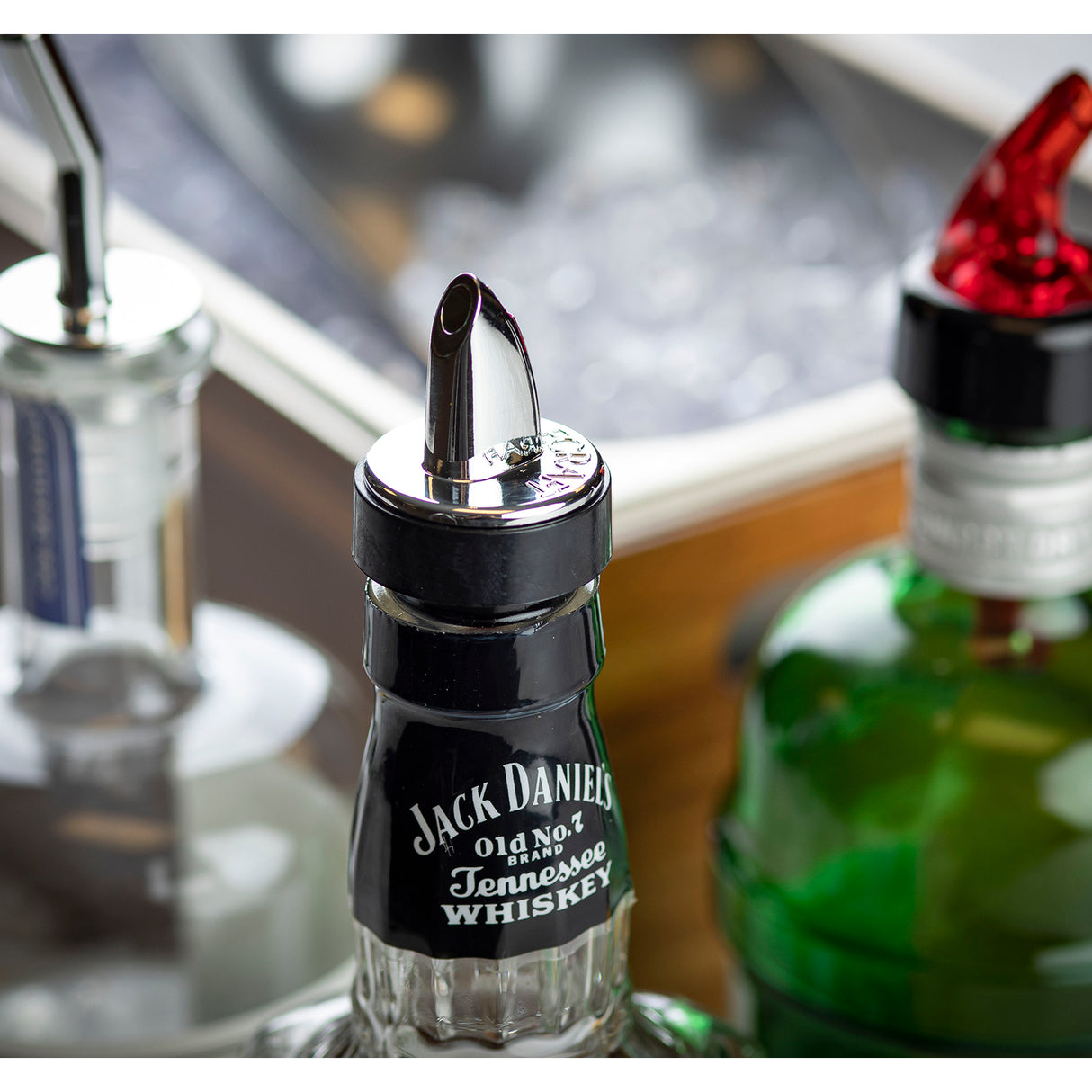 A close-up of a Jack Daniel’s Tennessee Whiskey bottle with a chrome plated spout. Nearby are part of a green bottle and clear bottle with pourers. In the blurred background, a metal container with ice evokes a classic bar setup.
