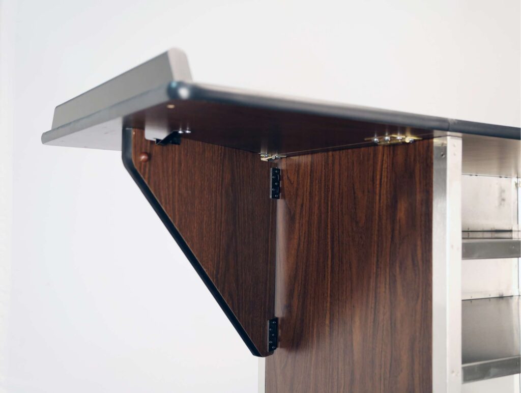 Close-up of a wooden countertop with a dark finish, attached to a stainless steel structure. A triangular wooden support bracket with visible hinges secures the drop leaves, and a black edge trim lines the top. The background is plain white.