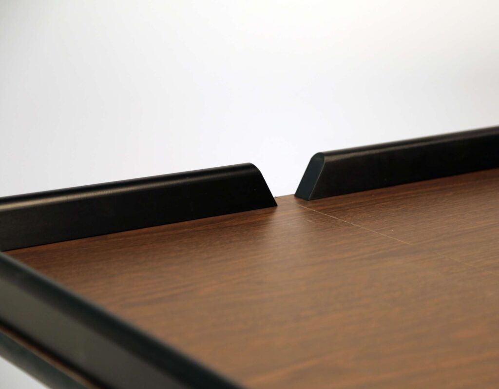 A close-up of a wooden tabletop with a smooth brown finish designed for a beverage service cart. Two black, angled, raised edges face each other near the center, leaving a narrow gap. The softly lit background highlights the table’s surface and details.