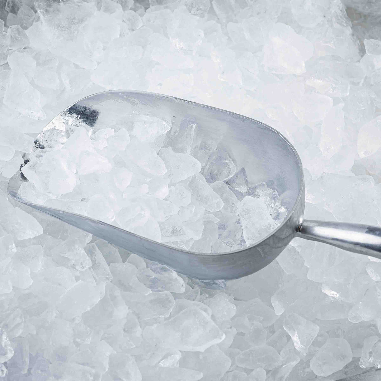 An aluminum Ice Scoop, 85 oz in size, is filled with small, clear ice cubes and rests atop a larger pile of ice. The sturdy scoop has a shiny, reflective surface and handle, enhancing the cold and frosty atmosphere of the scene.