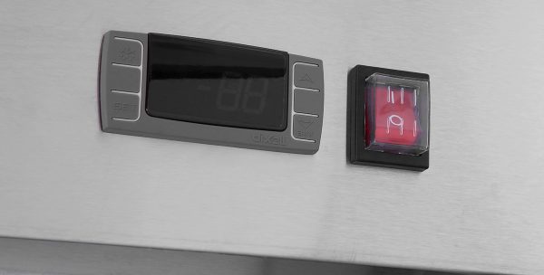 A digital control panel with a dark display and four buttons is mounted on metal. To its right is a red square switch with two white lines and a lock icon, indicating power and lock functions. The design is minimal, emphasizing function and clarity—hallmarks of the Atosa MBB69GGR Glass Door Refrigerator Back Bar Cooler.