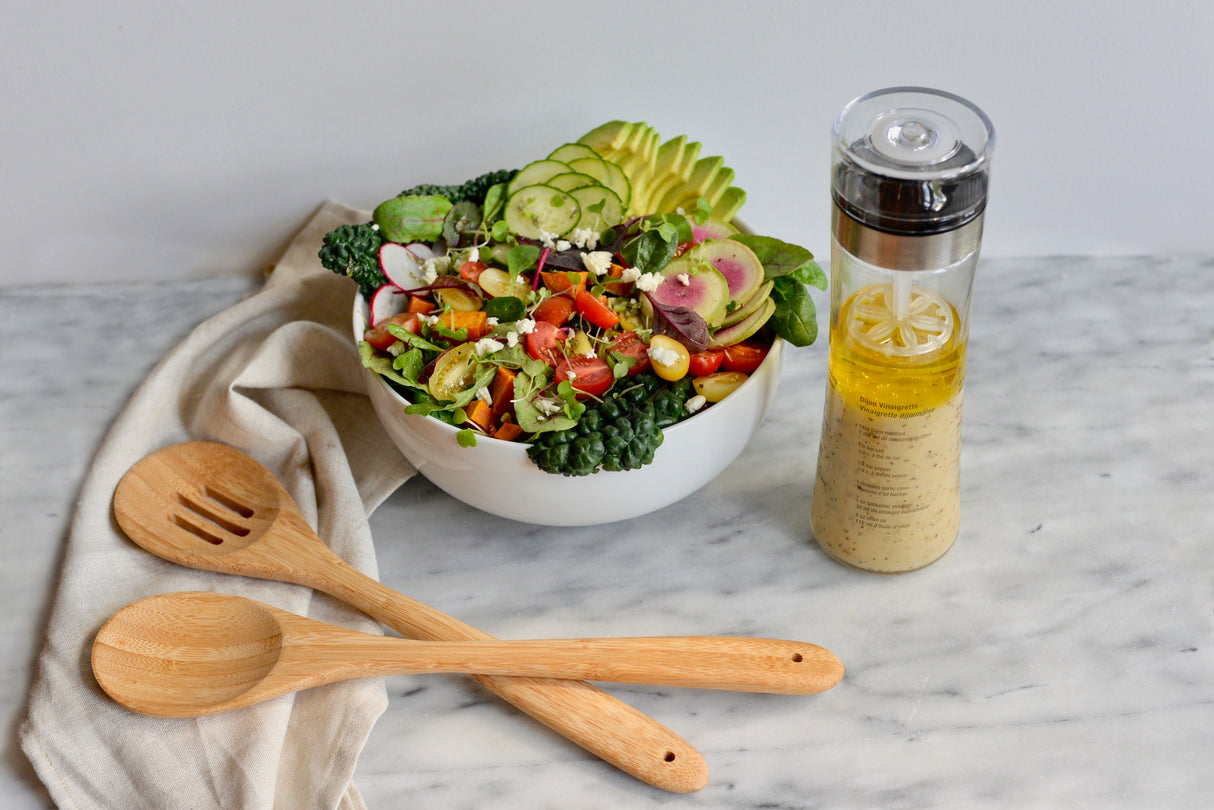 A colorful salad with avocado, radish, cucumber, cherry tomatoes, greens, and sprouts sits in a white bowl on a marble surface. Nearby are a beige cloth napkin, a 14 inch bamboo spoon, and a clear bottle of salad dressing with lemon.