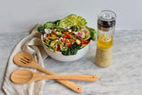 A colorful salad with avocado, radish, cucumber, cherry tomatoes, greens, and sprouts sits in a white bowl on a marble surface. Nearby are a beige cloth napkin, a 14 inch bamboo spoon, and a clear bottle of salad dressing with lemon.