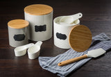 Three white and gray canisters with bamboo lids and blank black labels sit on a dark wood surface. Two canisters are open, showing white melamine measuring cups inside. A striped kitchen towel and a spatula with a wooden handle are beside the canisters.