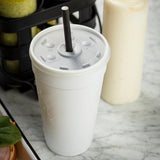 A white Styrofoam cup with a plastic lid and black straws stands on a marble countertop, condensation on its surface. Nearby are a squeeze bottle with light-colored sauce and a tray holding some green leafy vegetables.