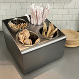 A stainless steel flatware cylinder holder contains wooden forks, spoons, knives, and striped paper straws. It sits on a gray countertop next to stacked round wooden plates, with a white subway tile backsplash in the background.