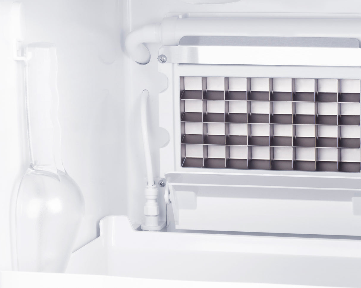 Close-up of a stainless steel icemaker inside a freezer, featuring a grid of square molds and metallic components. To the left, a frosted glass bottle stands upright against the white interior wall, highlighting clear ice production.