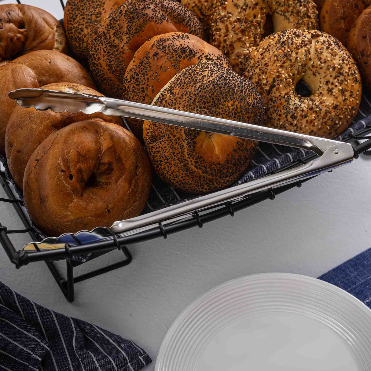 A wire basket lined with a dark striped cloth holds a variety of bagels, while stainless steel tongs rest on the basket. A stack of white plates sits nearby on the white surface, ready for serving.
