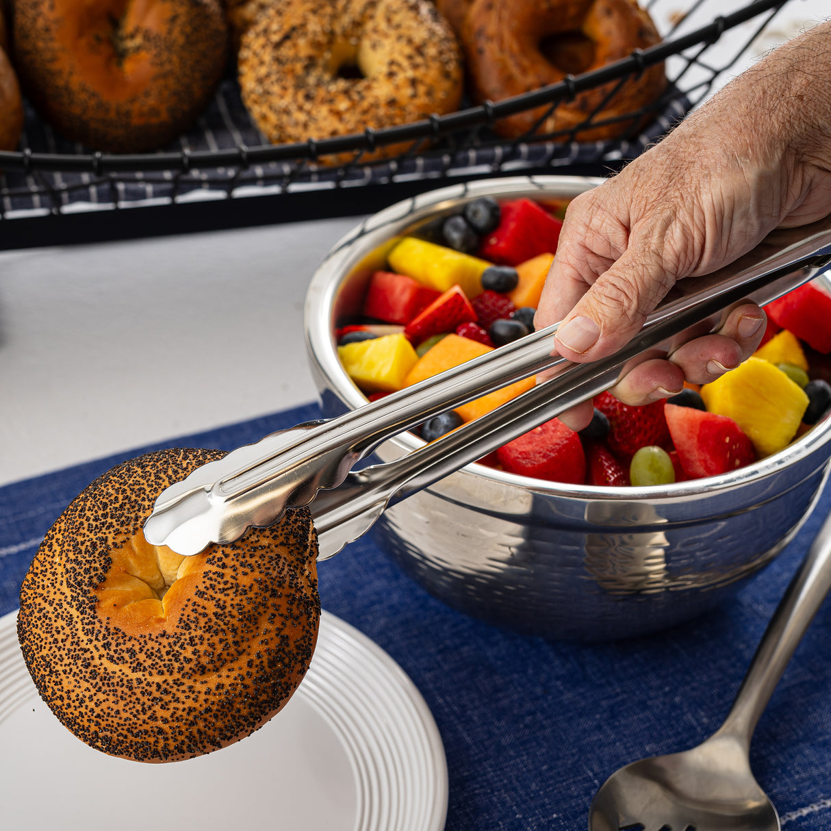 A hand uses stainless steel tongs to pick up a poppy seed bagel above a stack of white plates. In the background, there’s a shiny metal bowl of assorted fresh fruit and a basket of bagels. A large spoon rests on a blue napkin nearby.